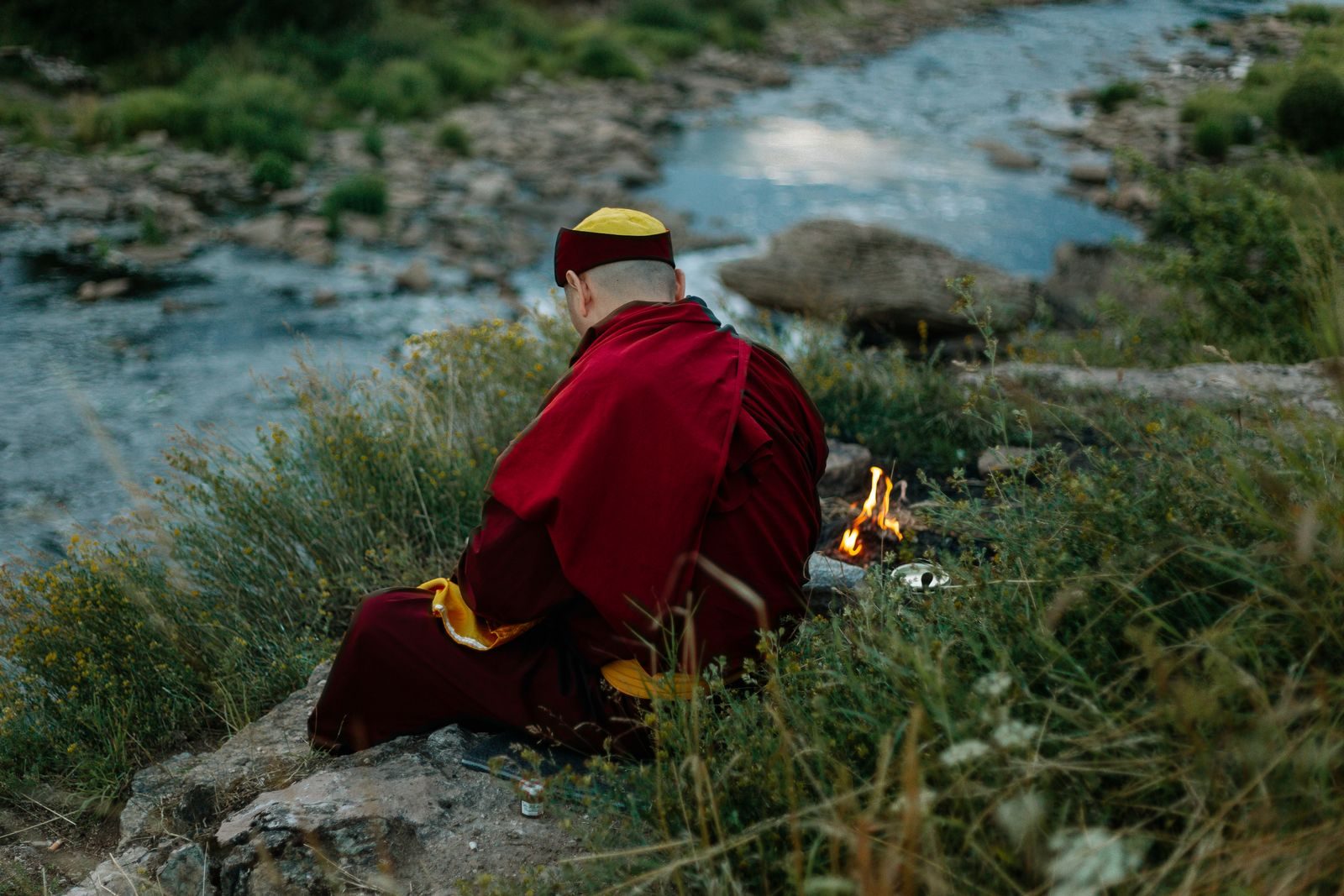 A robed figure tending a small fire by a stream